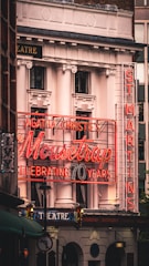 A theater building facade adorned with classic architectural elements features a prominent neon sign celebrating Agatha Christie's play 'The Mousetrap' marking its 70th anniversary. The sign in bright red lights is juxtaposed against the building's white columns and ornate decorations. Additional signage indicates the name 'ST MARTINS' vertically, contributing to the overall vintage charm.