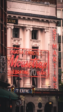 A theater building facade adorned with classic architectural elements features a prominent neon sign celebrating Agatha Christie's play 'The Mousetrap' marking its 70th anniversary. The sign in bright red lights is juxtaposed against the building's white columns and ornate decorations. Additional signage indicates the name 'ST MARTINS' vertically, contributing to the overall vintage charm.