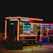 A food truck featuring neon lights displays a sign for fish and chips. The truck is parked in a dimly lit setting during the night. A person stands nearby, likely a customer.