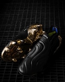 A pair of sleek black soccer cleats with a smooth, wavy design, featuring striking golden metal studs and detailing on the sole. The word 'COPA' is visible on the upper part of the shoe, and the cleats are placed on a dark, grid-like surface, enhancing their luxurious appearance.