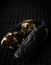 A pair of sleek black soccer cleats with a smooth, wavy design, featuring striking golden metal studs and detailing on the sole. The word 'COPA' is visible on the upper part of the shoe, and the cleats are placed on a dark, grid-like surface, enhancing their luxurious appearance.