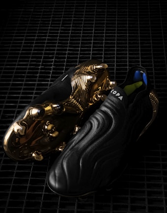 A pair of sleek black soccer cleats with a smooth, wavy design, featuring striking golden metal studs and detailing on the sole. The word 'COPA' is visible on the upper part of the shoe, and the cleats are placed on a dark, grid-like surface, enhancing their luxurious appearance.