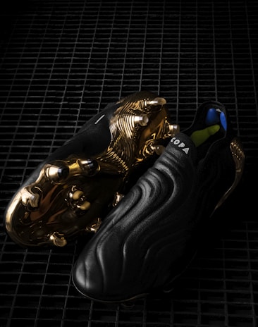 A pair of sleek black soccer cleats with a smooth, wavy design, featuring striking golden metal studs and detailing on the sole. The word 'COPA' is visible on the upper part of the shoe, and the cleats are placed on a dark, grid-like surface, enhancing their luxurious appearance.