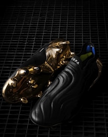 A pair of sleek black soccer cleats with a smooth, wavy design, featuring striking golden metal studs and detailing on the sole. The word 'COPA' is visible on the upper part of the shoe, and the cleats are placed on a dark, grid-like surface, enhancing their luxurious appearance.