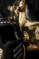 A close-up view of adidas soccer cleats with a striking black and gold color scheme. The soleplate features a unique gold design with cleat studs visible. The black upper part of the cleats has three white stripes and visible branding.