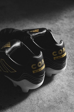 A pair of black soccer cleats with yellow accents, featuring the word 'Copa' and additional branding. The shoes are placed on a textured, gray surface and skillfully lit to create a moody atmosphere.