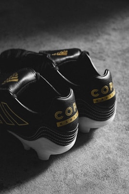 A pair of black soccer cleats with yellow accents, featuring the word 'Copa' and additional branding. The shoes are placed on a textured, gray surface and skillfully lit to create a moody atmosphere.