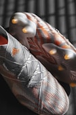 A pair of soccer cleats with a striped pattern in black and white, accented by orange details. The cleats are positioned on a dark surface with lighting that creates a grid-like shadow.