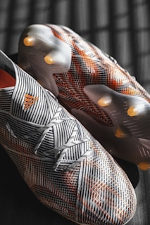 A pair of soccer cleats with a striped pattern in black and white, accented by orange details. The cleats are positioned on a dark surface with lighting that creates a grid-like shadow.