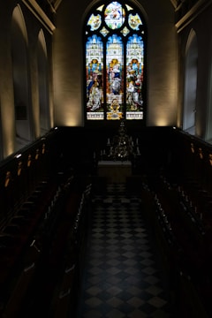 A dark, ornate chapel with a checkered floor and wooden pews arranged in rows. At the far end, there is a large, detailed stained glass window with colorful depictions of religious scenes. Beneath the window, there is an altar, and above the altar, a chandelier with candles.