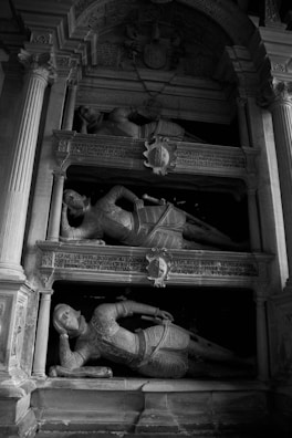 A monochrome image featuring a historical monument with detailed carvings and inscriptions. It displays three statues of knights in reclining positions, each wearing ornate armor. The structure is framed by classical architectural elements, including columns and arches.
