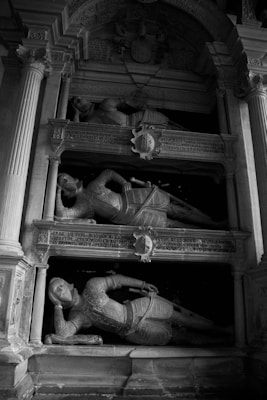 A monochrome image featuring a historical monument with detailed carvings and inscriptions. It displays three statues of knights in reclining positions, each wearing ornate armor. The structure is framed by classical architectural elements, including columns and arches.