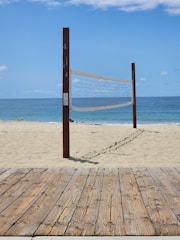 A sandy beach with a volleyball net set up between two wooden poles. The net is positioned parallel to the shoreline with the ocean in the background. A wooden boardwalk lies in the foreground.