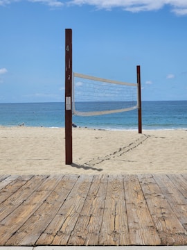 A sandy beach with a volleyball net set up between two wooden poles. The net is positioned parallel to the shoreline with the ocean in the background. A wooden boardwalk lies in the foreground.