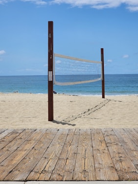 A sandy beach with a volleyball net set up between two wooden poles. The net is positioned parallel to the shoreline with the ocean in the background. A wooden boardwalk lies in the foreground.