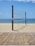 A sandy beach with a volleyball net set up between two wooden poles. The net is positioned parallel to the shoreline with the ocean in the background. A wooden boardwalk lies in the foreground.