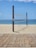 A sandy beach with a volleyball net set up between two wooden poles. The net is positioned parallel to the shoreline with the ocean in the background. A wooden boardwalk lies in the foreground.