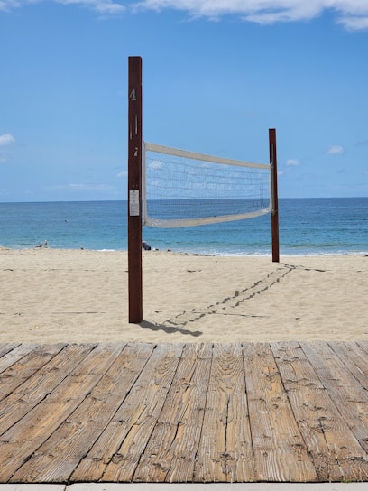 A sandy beach with a volleyball net set up between two wooden poles. The net is positioned parallel to the shoreline with the ocean in the background. A wooden boardwalk lies in the foreground.