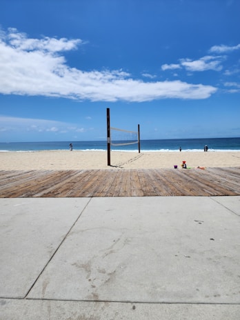 A beach scene on a sunny day shows a volleyball net set up on a sandy area with a pathway made of wood and concrete leading to the sand. In the background, a few people are near the shoreline where the ocean meets the beach. Some children's toys are scattered on the wooden deck.