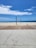 A beach scene on a sunny day shows a volleyball net set up on a sandy area with a pathway made of wood and concrete leading to the sand. In the background, a few people are near the shoreline where the ocean meets the beach. Some children's toys are scattered on the wooden deck.