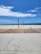 A beach scene on a sunny day shows a volleyball net set up on a sandy area with a pathway made of wood and concrete leading to the sand. In the background, a few people are near the shoreline where the ocean meets the beach. Some children's toys are scattered on the wooden deck.