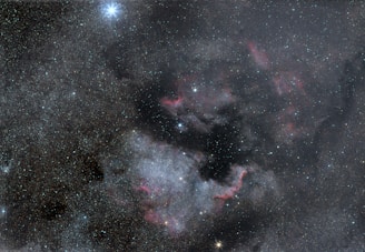 A vast view of space featuring countless stars scattered across the dark expanse. A prominent bright star stands out on the top left. Nebulous clouds with hints of red and pink hues swirl in the center, creating a captivating celestial formation.