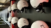 Several white helmets with numbers and logos are mounted in a neat row on a dark wall. The helmets have a sleek design, featuring brands like Audi, and are lit by a subtle red light creating a modern and organized appearance. A portion of a plant is visible on the lower left, adding a touch of natural contrast to the setting.