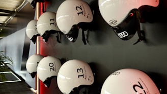Several white helmets with numbers and logos are mounted in a neat row on a dark wall. The helmets have a sleek design, featuring brands like Audi, and are lit by a subtle red light creating a modern and organized appearance. A portion of a plant is visible on the lower left, adding a touch of natural contrast to the setting.