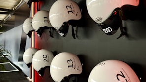 Several white helmets with numbers and logos are mounted in a neat row on a dark wall. The helmets have a sleek design, featuring brands like Audi, and are lit by a subtle red light creating a modern and organized appearance. A portion of a plant is visible on the lower left, adding a touch of natural contrast to the setting.