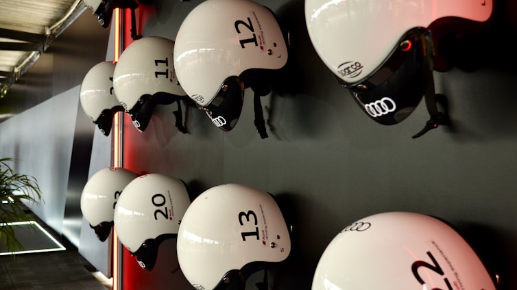 Several white helmets with numbers and logos are mounted in a neat row on a dark wall. The helmets have a sleek design, featuring brands like Audi, and are lit by a subtle red light creating a modern and organized appearance. A portion of a plant is visible on the lower left, adding a touch of natural contrast to the setting.