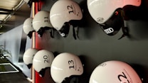 Several white helmets with numbers and logos are mounted in a neat row on a dark wall. The helmets have a sleek design, featuring brands like Audi, and are lit by a subtle red light creating a modern and organized appearance. A portion of a plant is visible on the lower left, adding a touch of natural contrast to the setting.