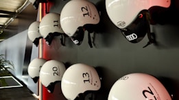 Several white helmets with numbers and logos are mounted in a neat row on a dark wall. The helmets have a sleek design, featuring brands like Audi, and are lit by a subtle red light creating a modern and organized appearance. A portion of a plant is visible on the lower left, adding a touch of natural contrast to the setting.