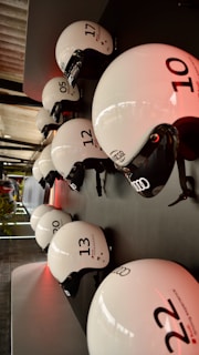 Multiple white helmets with numbers and branding are neatly arranged in two rows on a dark surface. The lighting is warm, and the helmets have a glossy finish that reflects the surrounding light. Visible in the background are some structural elements and a hint of greenery.