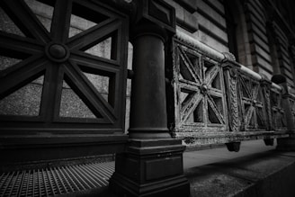A close-up perspective of a wrought iron fence with ornate, geometric patterns each featuring multiple intersecting lines and circular central motifs. The metal appears weathered, showing signs of rust and wear, positioned in front of a stone wall with a textured surface. The scene is captured in a muted color palette with a focus on architectural detail.