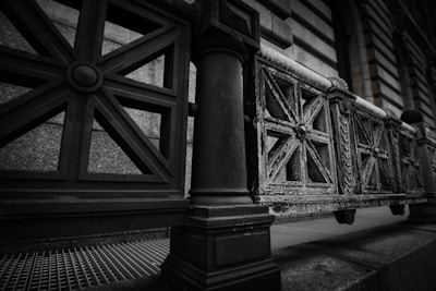 A close-up perspective of a wrought iron fence with ornate, geometric patterns each featuring multiple intersecting lines and circular central motifs. The metal appears weathered, showing signs of rust and wear, positioned in front of a stone wall with a textured surface. The scene is captured in a muted color palette with a focus on architectural detail.