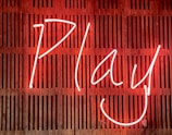 Bright neon sign displaying the word 'Play' with a smiling face, set against a wooden slatted background.