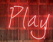 Bright neon sign displaying the word 'Play' with a smiling face, set against a wooden slatted background.