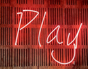 Bright neon sign displaying the word 'Play' with a smiling face, set against a wooden slatted background.