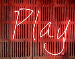 Bright neon sign displaying the word 'Play' with a smiling face, set against a wooden slatted background.