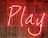 Bright neon sign displaying the word 'Play' with a smiling face, set against a wooden slatted background.