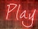 Bright neon sign displaying the word 'Play' with a smiling face, set against a wooden slatted background.