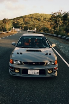 A silver sports car is parked on an empty road surrounded by lush greenery and hills. The car has a distinctive front design with round yellow fog lights and a Japanese license plate. The road is flanked by guardrails, leading into a background of dense, green trees under a partly cloudy sky.