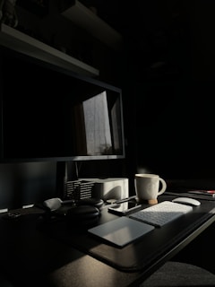 A dimly lit workspace with a computer monitor, keyboard, mouse, and various office items on a desk. A coffee mug is positioned to the right of the keyboard. Natural light from a window casts shadows across the room, highlighting the clean, organized setup.