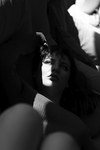 A person is lying down, partially illuminated by a beam of light that casts dynamic shadows across their face and body, creating a dramatic contrast between light and darkness. The expression on their face appears thoughtful or introspective.