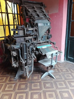 A vintage linotype machine is positioned in a room with patterned tile flooring and large windows. The machine is complex, with numerous metal components, levers, and knobs, and a keyboard at the base. The room's walls are painted in a muted pink shade, complementing the industrial aesthetic of the machine.