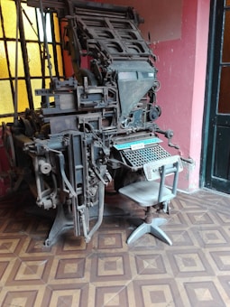 A vintage linotype machine is positioned in a room with patterned tile flooring and large windows. The machine is complex, with numerous metal components, levers, and knobs, and a keyboard at the base. The room's walls are painted in a muted pink shade, complementing the industrial aesthetic of the machine.