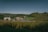 A sprawling landscape featuring rolling hills covered with fields and trees. In the foreground, there is a large field of crops, possibly corn, leading to a cluster of industrial buildings with metal silos and storage facilities. In the background, more agricultural fields and patches of forest extend towards the horizon. The sky is clear with a few scattered clouds, suggesting a calm and serene setting.