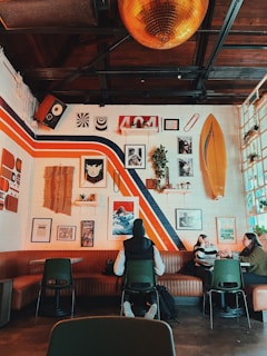 A cozy cafe interior features a mix of vintage and modern decor, with colorful wall art and a surfboard mounted on a brick wall. The ceiling has wood beams, a large golden disco ball, and plants adding a touch of greenery. Patrons, dressed casually, sit at tables against a brown leather booth, engaged in conversation.