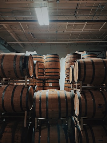 Large wooden barrels stacked in a warehouse, illuminated by overhead fluorescent lights. The barrels, likely used for aging whiskey or wine, are organized in rows with metal bands and wooden braces.