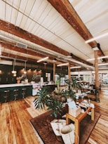 A cozy and rustic interior featuring wooden beams and floors. The space includes several tables displaying assorted products, plants, and decor items. The area is well-lit with natural tones and a warm atmosphere. A counter with stools is visible in the background, along with a sign reading 'Jones General'.